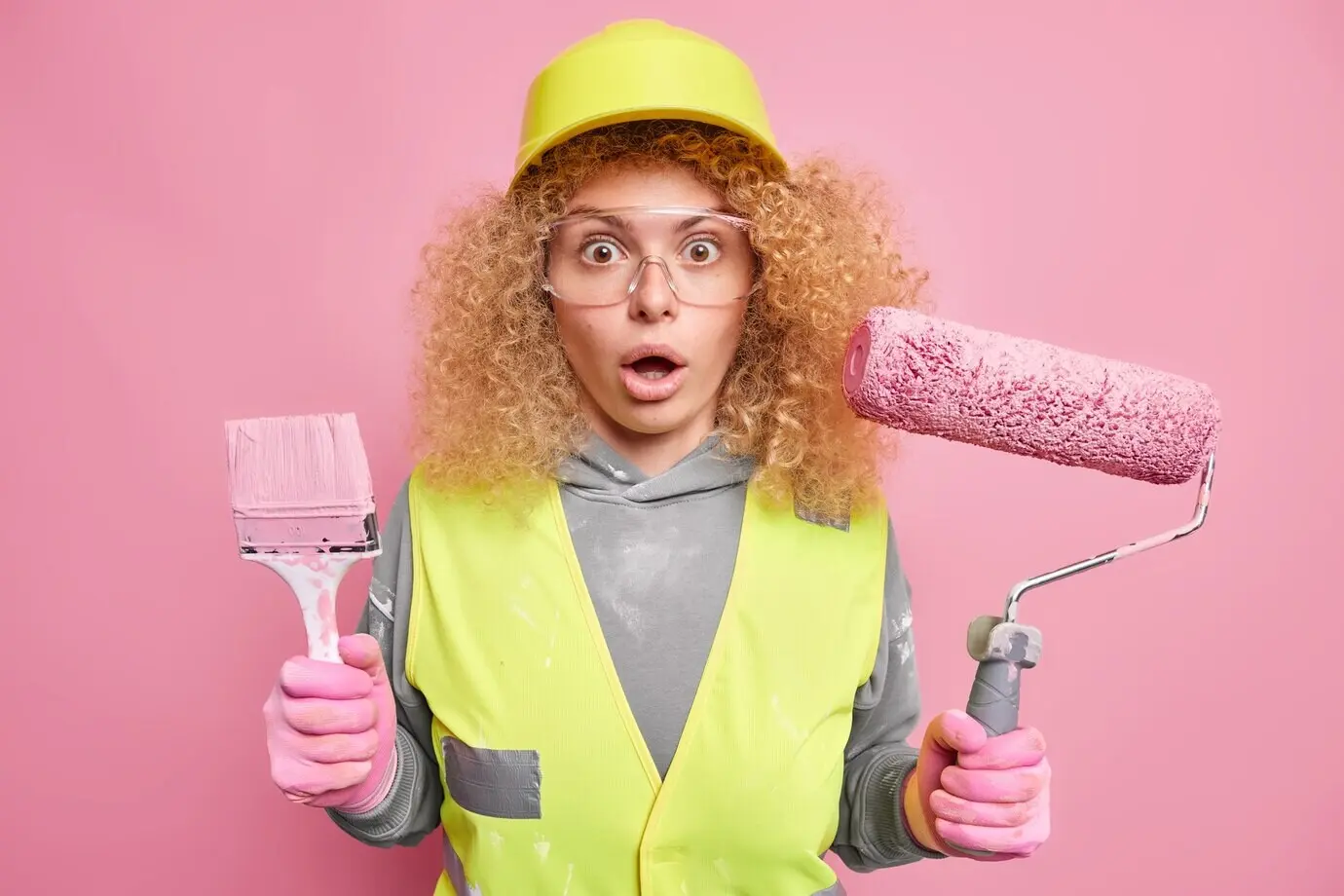 Eine Handwerkerin hält Malerpinsel und Farbrolle, reagiert mit erstauntem Gesichtsausdruck auf etwas Unglaubliches und ist als beschäftigte Arbeiterin in Uniform gekleidet.