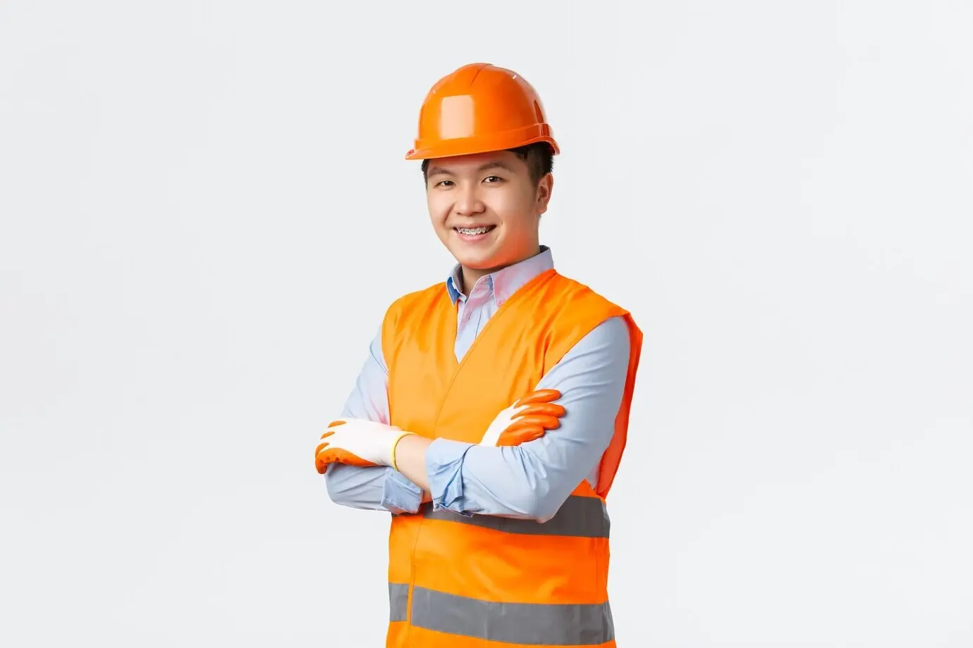 Konzept für Bauwesen und Industriearbeiter: Selbstbewusster junger asiatischer Ingenieur und Bauleiter in reflektierender Kleidung und mit Helm, mit verschränkten Armen und frechem Lächeln, stellt Qualität sicher, weiße Wand.